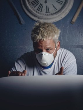 Atölyede gözlemlerken ve dokunurken beyaz sörf tahtasını yapan ve kontrol eden konsantre sarı saçlı bir erkek.