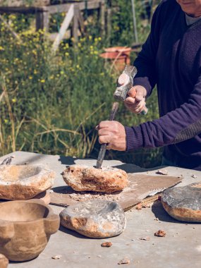 İsimsiz yaşlı bir zanaatkar çekiçle kaya yontarken açık hava atölyesinde taş kasesi yapıyor.