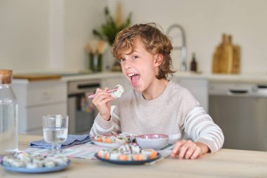 Evde yemek odasında pembe çubuklarla lezzetli suşi ruloları yerken kameraya bakıp eğlenen komik çocuk.