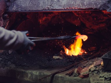 Kimliği belirsiz erkek nalbant eldiven ve maşayla demir ocağı atölyesinde çalışırken kızgın fırında metal yakıyor.