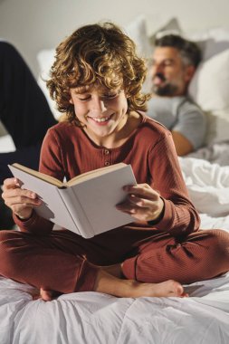 Pijamalı gülümseyen çocuk, evde babamın yumuşak yatağında yatmasına karşı ilginç bir kitap okurken eğleniyor.