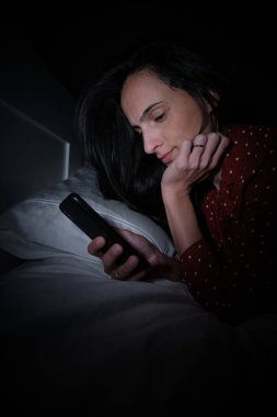 Gece geç saatlerde karanlık yatak odasında cep telefonundan mesajları kontrol ederken uykusuzluktan muzdarip İspanyol kadının yan görüntüsü