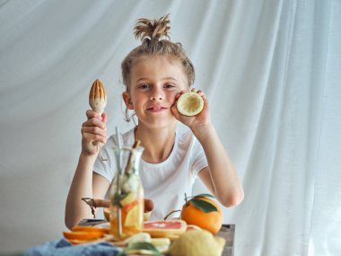Manüel ahşap meyve sıkacağı ve sıkılmış limonla masada meyve ve detoks suyuyla beyaz arka planda otururken kameraya bakan sevimli çocuk.