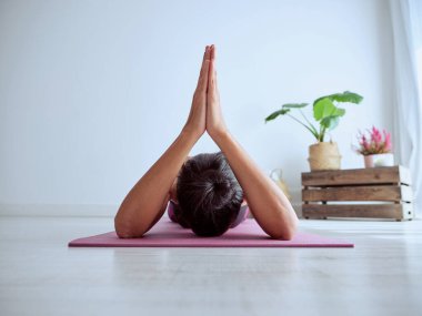 İsimsiz bir kadın, yeşil saksı bitkisiyle oturma odasında yoga yaparken elleri bağlı bir şekilde mindere uzanıyor.