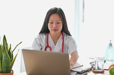 Modern netbook 'a göz atarken çağdaş hastanede çalışırken belgelerle masada oturan konsantre Asyalı kadın terapist.