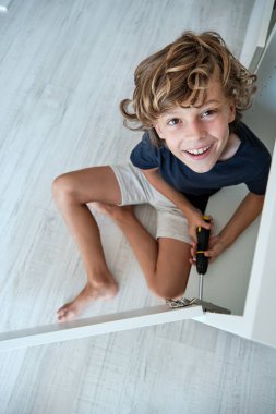 Yukardan gelen çıplak ayaklı, yerli giysili, çıplak ayaklı, tornavidayla ahşap mobilya monte eden bir çocuk.