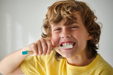 Kahverengi dalgalı sarı saçlı çocuk kameraya bakarken diş fırçasıyla diş fırçası takıyor.