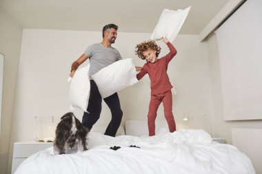 Yastıklarla yatağa atlayıp köpeklerle oynayan pozitif baba ve oğuldan oluşan bir vücut.