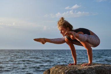 Çıplak ayaklı sporcu kadın sporcu, Firefly pozu veriyor. Kayalık sahilde yoga yaparken deniz kenarında sallanıyor.