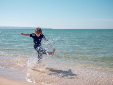Yaz mevsiminde ıslak sahilde oynarken durgun dalgalanan denizin berrak turkuaz suyunu sıçratan küçük bir çocuğun tüm vücudu.