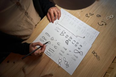 Kimliği belirsiz kuyumcunun üst görüntüsü. Tahta masa üzerine kalem ve mücevher çizimleri.