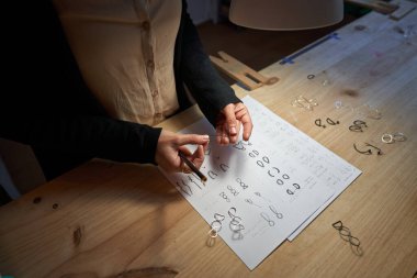 Lambanın altındaki masada takılarla çalışırken kağıt üzerine kalem çizen yüksek açılı isimsiz bir zanaatkar.