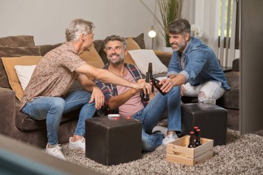 Orta yaşlı baba ve oğulların evde dinlenirken bira şişelerini tokuşturarak gülüşleri.