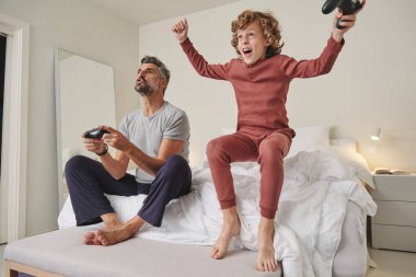 Bütün vücudu pijamalı, yatakta zıplayan, sakallı ve video oyunu oynayan neşeli bir çocuk.