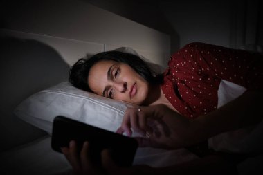 Gece geç saatlerde yatak odasında rahat bir yatakta yatarken modern cep telefonuna mesaj atan uyurgezer İspanyol kadın.