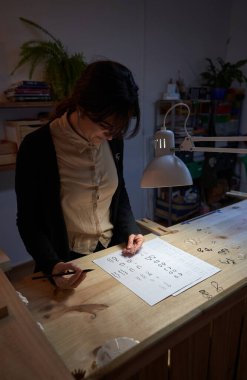 Odaklanmış kadın kuyumcunun yüksek açılı resmi kağıda çizerken parlayan lambanın yanında duruyordu.