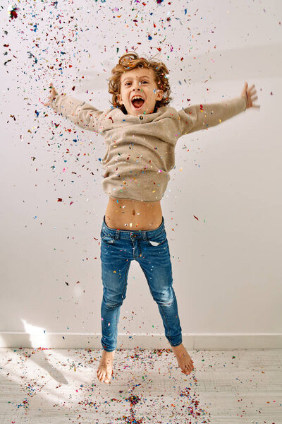 Full body of cheerful preschool boy in jeans and jumper jumping with raised hands and screaming while colorful confetti of cracker falling