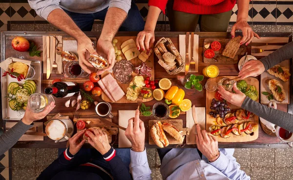 Top view of crop anonymous people eating tasty food while sitting at wooden table with assorted appetizers in light kitchen