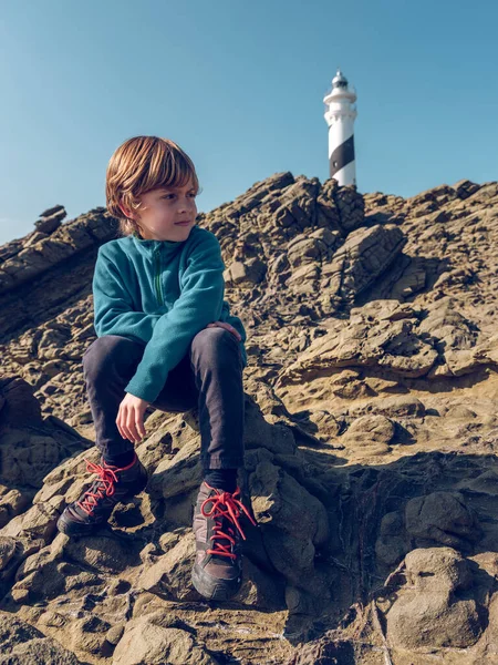 岩の上に座って白い灯台と雲のない青空を眺めているアクティブウェアの小さな男の子の全身 — ストック写真