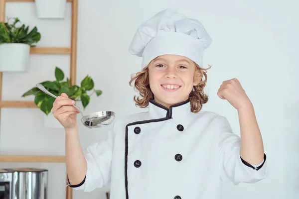 快乐地微笑着的小男孩 一头卷曲的金发 头戴白色厨师帽 身穿夹克衫 手拿着勺子站在厨房里 一边在家里做健康食品 一边看着相机 — 图库照片