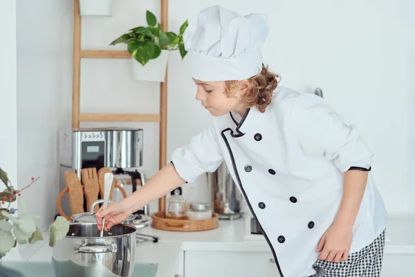 在家里厨房的炉灶上做饭时 穿着白色厨师制服的小孩用勺子搅拌锅里的食物的侧视图 — 图库照片