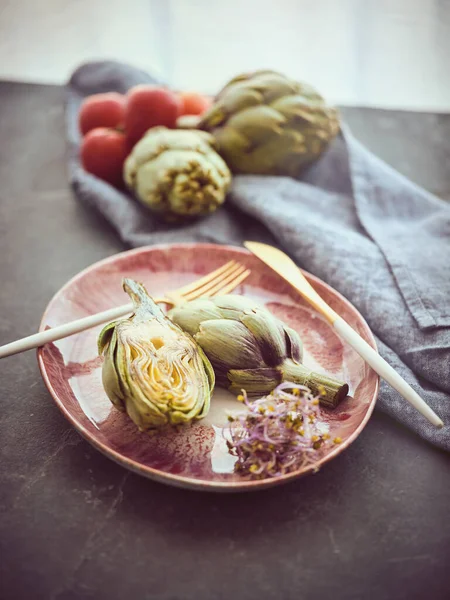 キッチンで熟したトマトの近くのテーブルの上にカトラリー付きのプレートにハーブ付きの新鮮なカットアーティチョークの上から — ストック写真