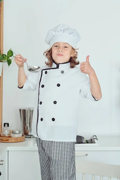 穿着白色厨师制服的积极的孩子 一边在厨房做饭 一边看着相机 一边用勺子尝着食物 并把大拇指伸出来 — 图库照片