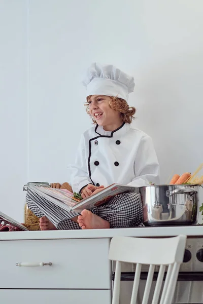 从下面看身穿厨师制服 头戴帽子 双腿交叉坐在厨房柜台上 打开菜谱的青春期男孩 — 图库照片