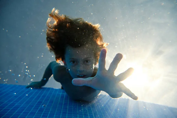 週末の夏の日に青いプールで泳いでいる間にカメラを見て手を差し伸べ空気の泡で囲まれた驚きの子供の下から — ストック写真