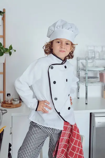Confident little kid in chef hat and jacket with grin standing with hands on waist while preparing food at home kitchen and looking at camera