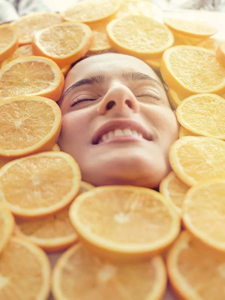 Face of positive woman lying in big heap of slices of juicy oranges with eyes closed