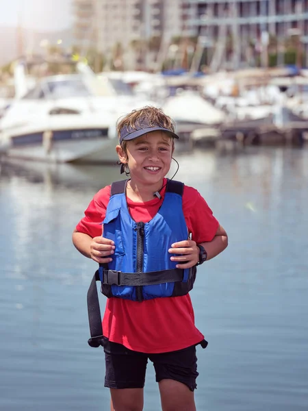 カメラを見ながら港近くに立っている安全装備と帽子を身に着けているポジティブな少年 — ストック写真