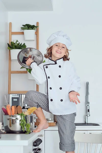 穿着厨师帽和西服 面带微笑的孩子站在锅盖的火炉边 一边在厨房里做饭 一边回头看 — 图库照片