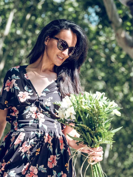黒い夏のドレスと公園のぼやけた背景に対する新鮮な白い花の束を身に着けている長い髪の魅力的なブルネットの下から — ストック写真