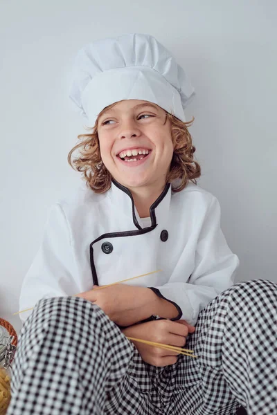 快乐地微笑着 穿着厨师制服 头戴帽子 可爱的十多岁男孩坐在厨房柜台上 望着远方 — 图库照片