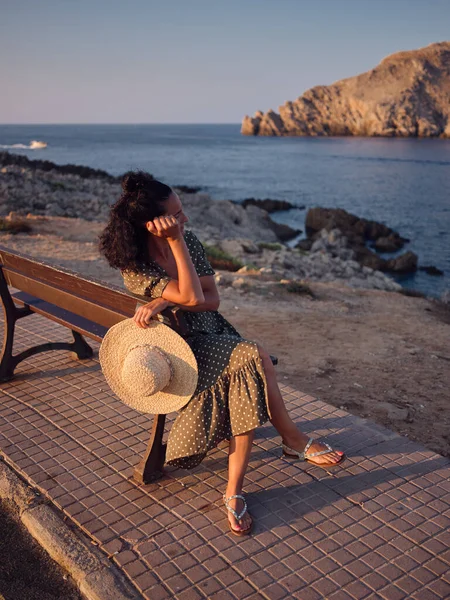 穿着时髦服装的女人坐在靠近大海的长椅上 在日落时享受暑假 同时靠着手 望着远方 — 图库照片