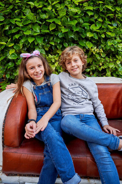 Happy preteen boy and girl in casual clothes smiling and looking at camera while sitting on old leather couch near green bushes in garden