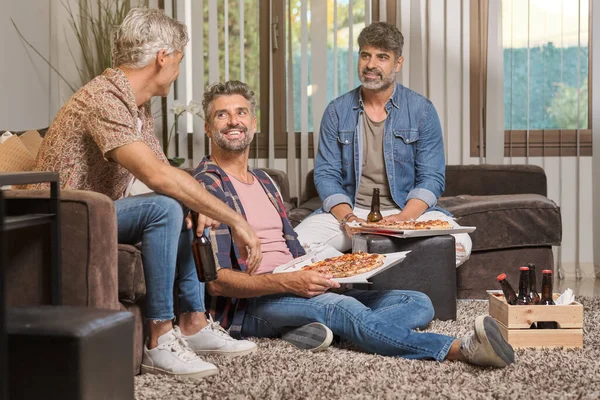 stock image Content adult men in casual clothes smiling and looking at each other while eating pizza and drinking beer in cozy living room