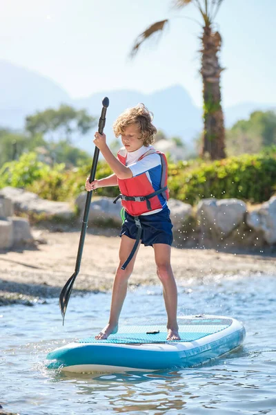 青いSupボードの上に立つ水着とライフジャケットに集中した子供の完全な長さ夏休みの間 砂浜の近くのパドルで漕ぐ — ストック写真