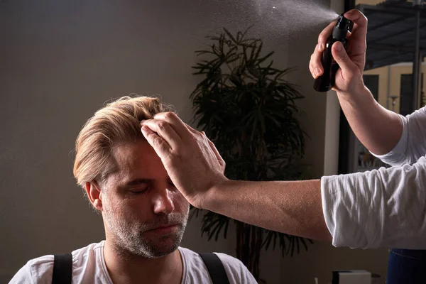 Mature man with eyes closed and unrecognizable barber working with hair of client with spray in barbershop