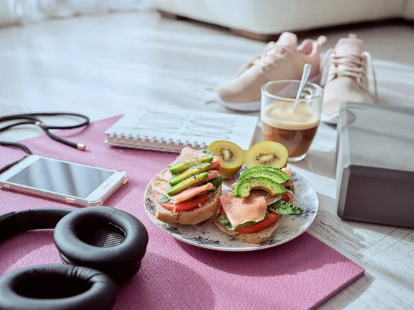 在家里的地板上 不同的运动器材旁边 有许多美味的烤面包 上面有水果和蔬菜 还有放在瑜伽垫上的咖啡 — 图库照片