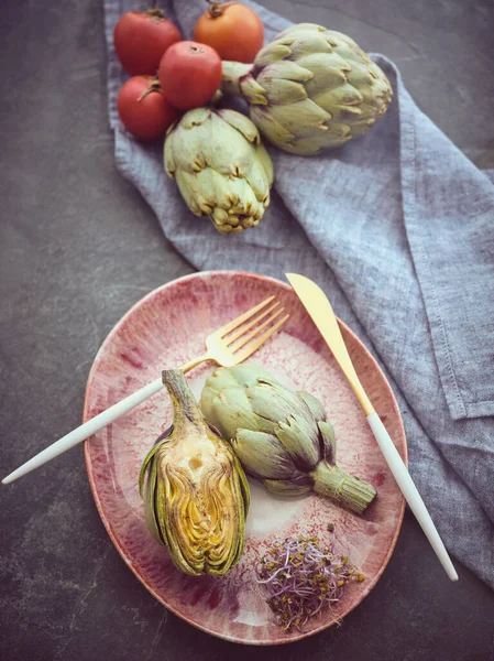 From above of fresh cut artichokes with herbs served on plate with cutlery on table near ripe tomatoes in kitchen
