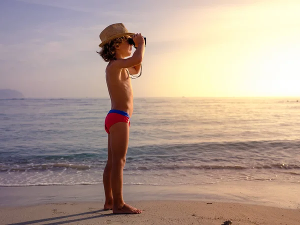 Hasır şapkalı, ıslak kumlu sahilde duran ve gün batımında dürbünle deniz manzarasının tadını çıkaran bir çocuk.