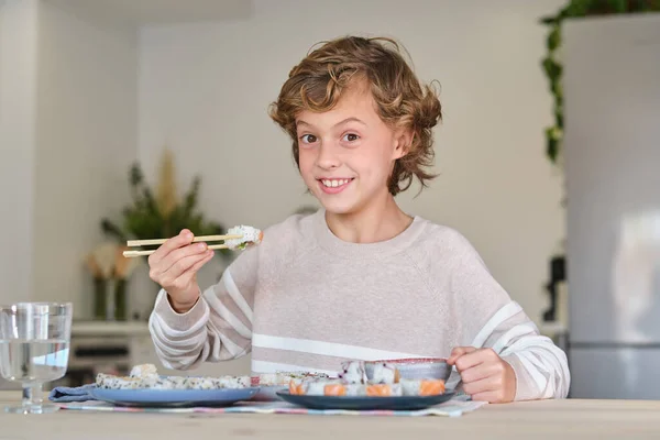 穿着休闲装的积极男孩笑着看着镜头 坐在桌旁 用筷子吃着面包卷 — 图库照片