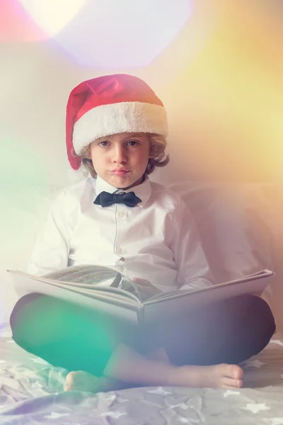 圣诞庆典期间 穿着西服 头戴圣诞礼帽的光着脚的男孩在床上看那本不感兴趣的书 一边看着相机 — 图库照片