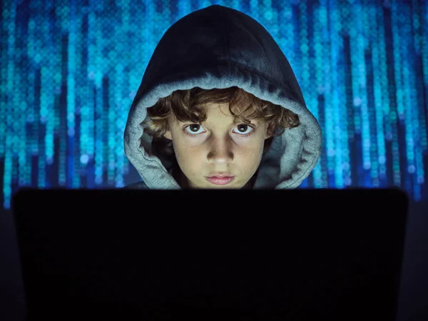 Bilgisayarlı ciddi bilgisayar korsanı çocuk karanlık ışıkta bulanık kod arka planında kameraya bakıyor.