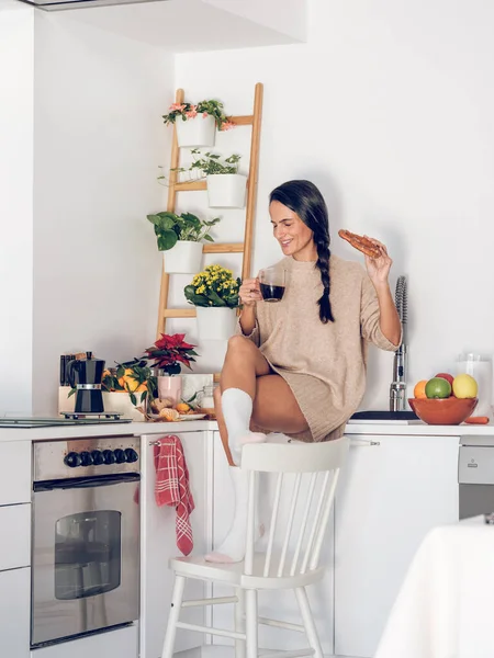 Full body of happy ethnic woman with long black hair sitting on kitchen counter and drinking coffee while enjoying breakfast at home in morning