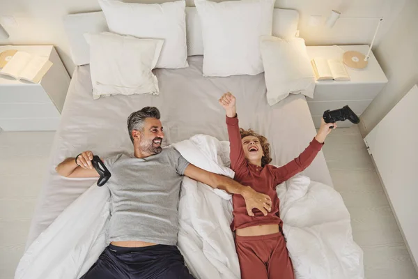 stock image From above of laughing man tickling boy while lying on bed with joystick and having fun