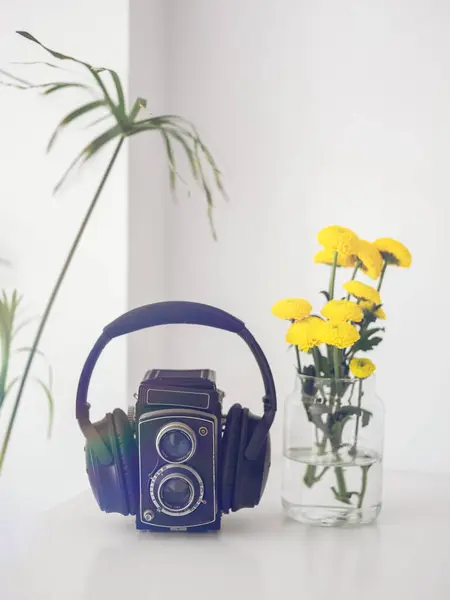 Musical headphones on vintage black photo camera placed on white table near vase with yellow flowers in room at home