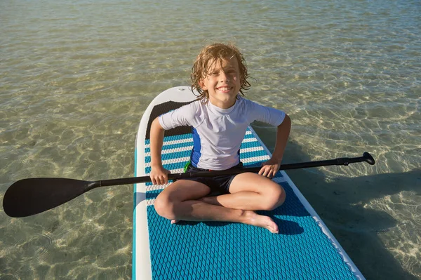 フルボディポートレートの笑顔の男の子とパドルで座っていますSupボードと夏休み中に海の真ん中にカメラを見て — ストック写真